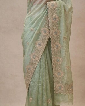 A premium, flat-lay view of a hand-embroidered mint green silk chiffon saree with delicate silver embellishments, paired with a matching blouse piece.