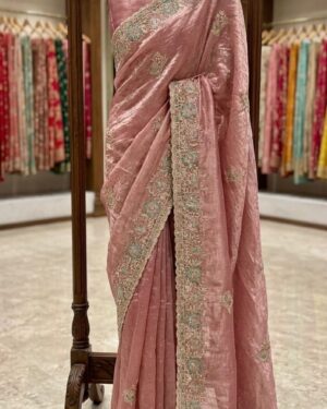 A premium rose gold silk saree with intricate floral embroidery and detailed borders, elegantly displayed on a wooden hanger stand in a boutique setting.