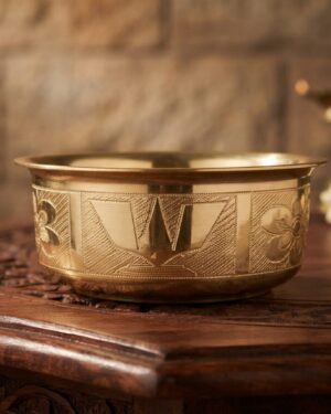 A small, polished brass prasadham bowl featuring intricate hand-etched floral patterns and a central Venkateshuni Namalu engraving, set on a dark wooden table in a traditional puja setting.