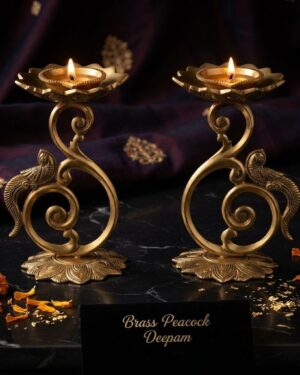 A pair of premium handcrafted brass peacock deepams with lotus bases and swirl stands, placed on a dark surface decorated with marigold petals and subtle gold accents against a rich purple fabric background.