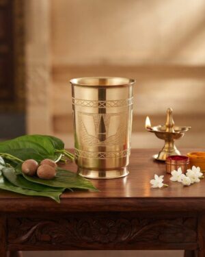 A small, polished brass tumbler displayed on a white surface, featuring detailed hand-etched geometric borders and a prominent Venkateshuni Namalu engraving at the center.