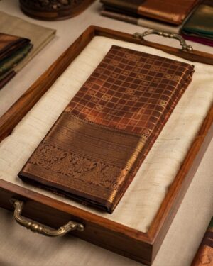 A chocolate brown Exquisite Rust Colored Semi-Pattu Sareeand an intricate antique gold zari border, neatly folded and displayed in a rustic wooden tray.