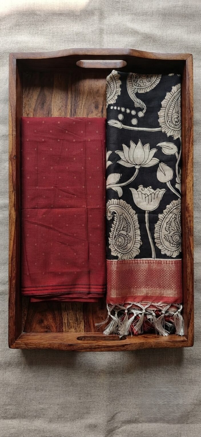 Unstitched maroon Pochampalli Ikat top fabric next to a black and cream handcrafted floral Kalamkari dupatta, neatly folded in a decorative wooden tray.