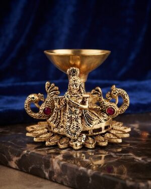 Close-up shot of a polished brass Krishna diya featuring peacock motifs and red gemstone accents against a rich blue velvet and marble background
