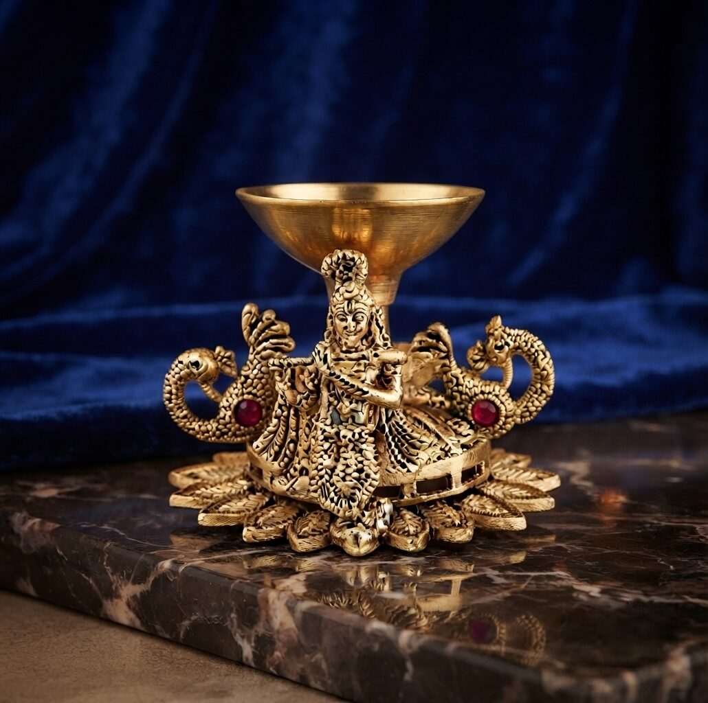 Close-up shot of a polished brass Krishna diya featuring peacock motifs and red gemstone accents against a rich blue velvet and marble background