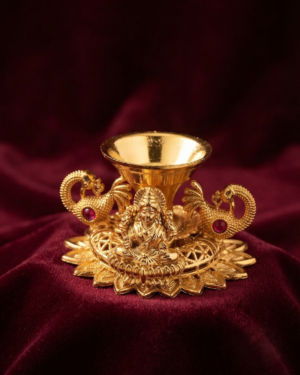 An Exquisite Gold Lakshmi Diya oil lamp featuring sculpted peacock handles and a Goddess Lakshmi motif, displayed on a deep maroon velvet fabric.