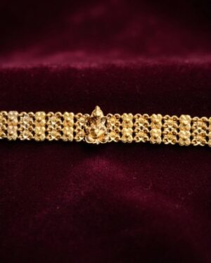 Close-up of an intricate matte gold Rakhee bracelet featuring a central Ganesha figurine and multi-row floral link patterns on a rich black textured surface.