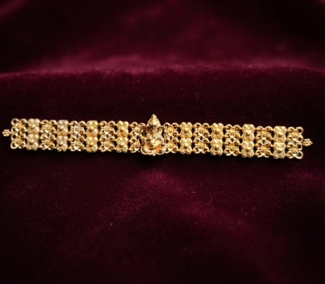 Close-up of an intricate matte gold Rakhee bracelet featuring a central Ganesha figurine and multi-row floral link patterns on a rich black textured surface.