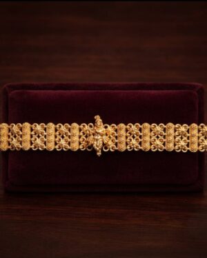 Elegant studio shot of a traditional matte gold Rakhee bracelet highlighting a divine Krishna centerpiece and delicate filigree beadwork against a deep burgundy velvet background.
